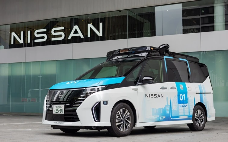 日産本社ビルの前に停車する「日産セレナをベースとした自動運転車両」。車両は左前方を向いており、白を基調とした車体に青と黒の配色が施されている。車両上部には自動運転関連の機器が搭載されている。背景には日産本社ビルの外観とNISSANのロゴが見え、屋外の明るい空間で撮影された車両の様子を示している。