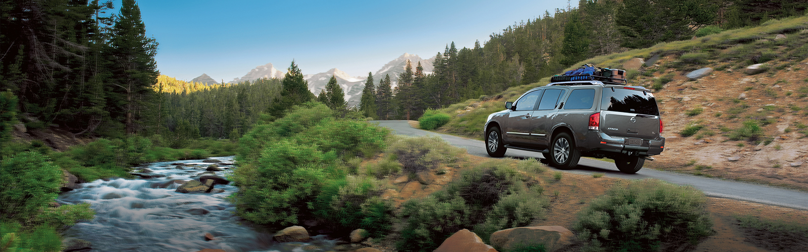 The back view for the Nissan Armada driving around a curve with a creek to the left and tall trees in the background.