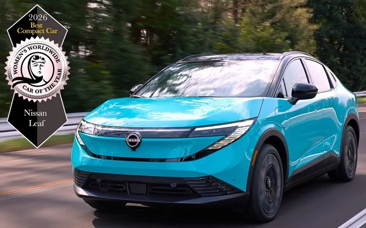 A pale blue Nissan LEAF, visible from the front three-quarter perspective drives along the road. The image includes the logo of the Womens’ Worldwide Car Of The Year logo - best compact car 2026