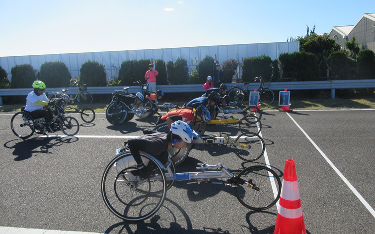 At Nissan Oppama Plant GRANDRIVE outdoor track, several racing wheelchairs are lined up with participants wearing helmets in a forward start position. The background shows green hedges, white buildings, and a clear blue sky, with a red traffic cone placed in the foreground.