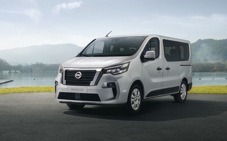 Ein silberner Nissan Primastar Kombi steht auf einem asphaltierten Platz neben einer Wiese und einem See, 3/4 Frontansicht