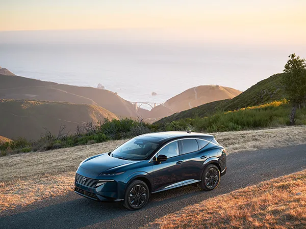 A blue 2025 Nissan Kicks parked in front of a body of water with a bridge and trees in the background