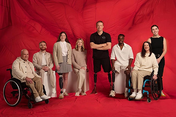 A group of eight people posed together against a red studio background. The group includes single and double-leg amputees, wearing prosthetic legs, as well as two people in wheelchairs. Paralympic athlete Richard Whitehead MBE stands at the centre of the group.