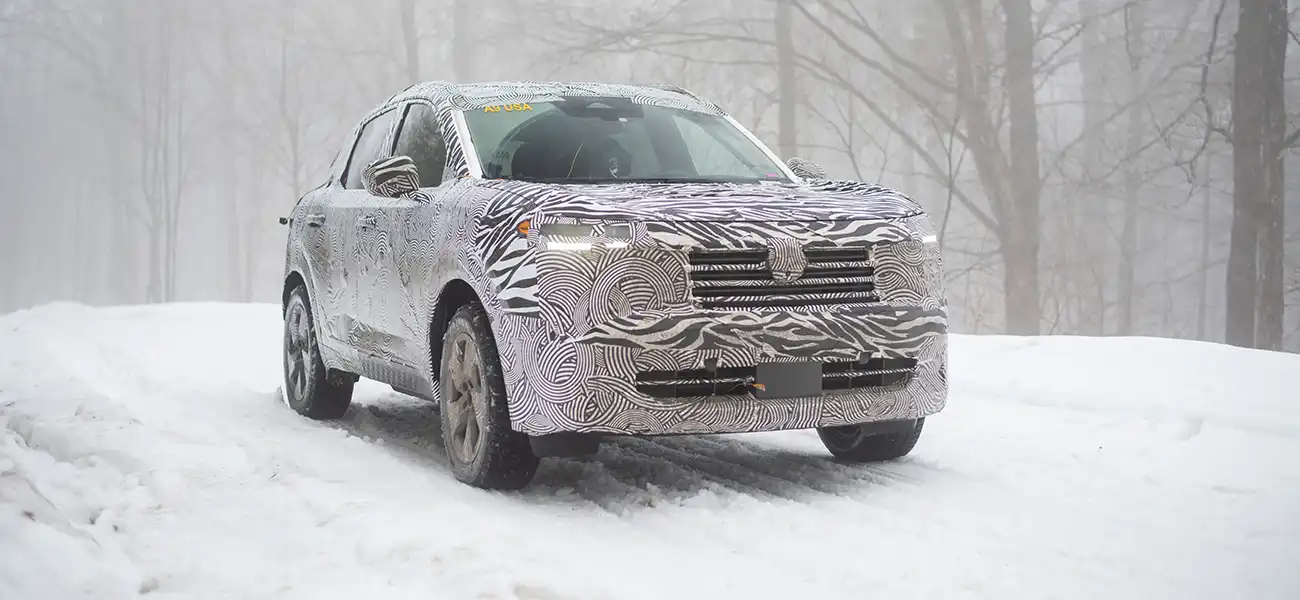 A white camouflaged 2025 Nissan Kicks driving along a snowy path in the woods.