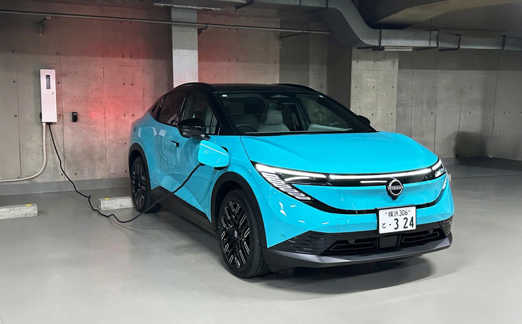 地下駐車場で、ターコイズブルーの車体の日産リーフが左前方から見える向きで駐車し、左側面から延びたケーブルを壁の充電器につないで充電している。
