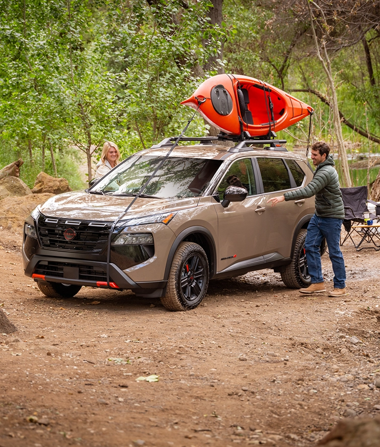 Front left 3/4 angle of a 2026 Nissan Rogue parked on a camp site with a kayak on its roof and a man opening the door.