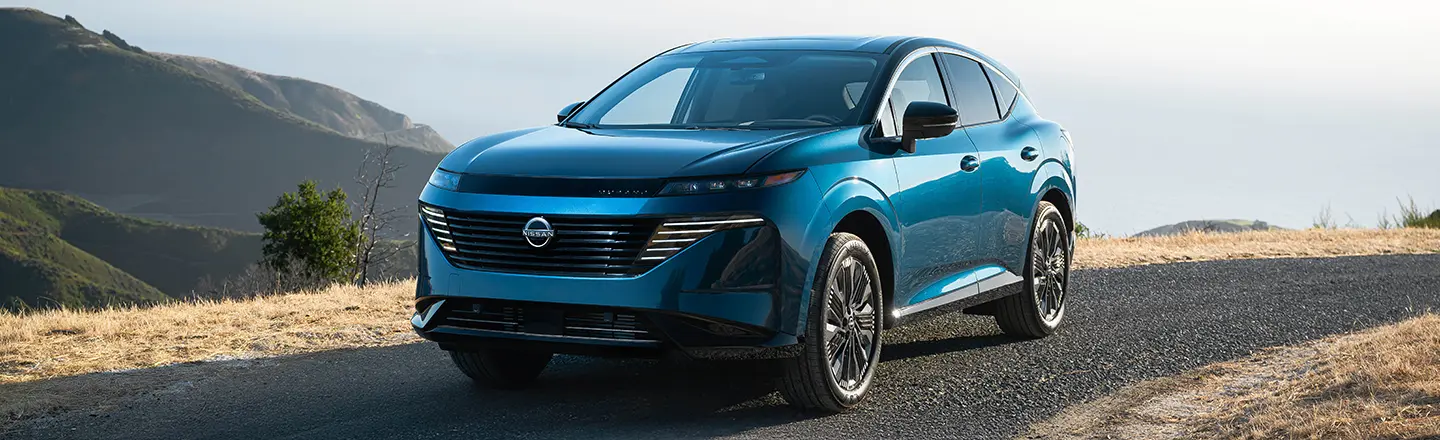 Front left 3/4 view of a blue 2025 Nissan Murano parked on a hill overlooking the ocean with other hills beside.