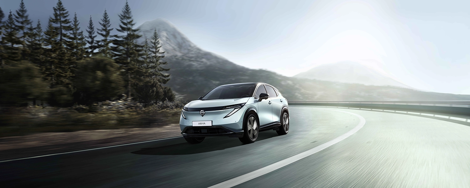 Nissan Ariya Model Year 2026 driving on a forest bordered route, emphasizing the coupe inspired SUV profile.