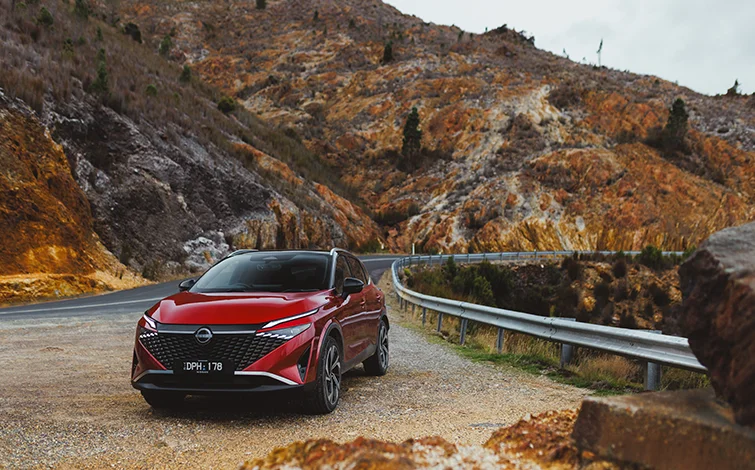 2026 Nissan Qashqai Ti-L E-POWER finished in Fuji Red with Black Roof parked beside a winding mountain road with rugged rock formations in the background.