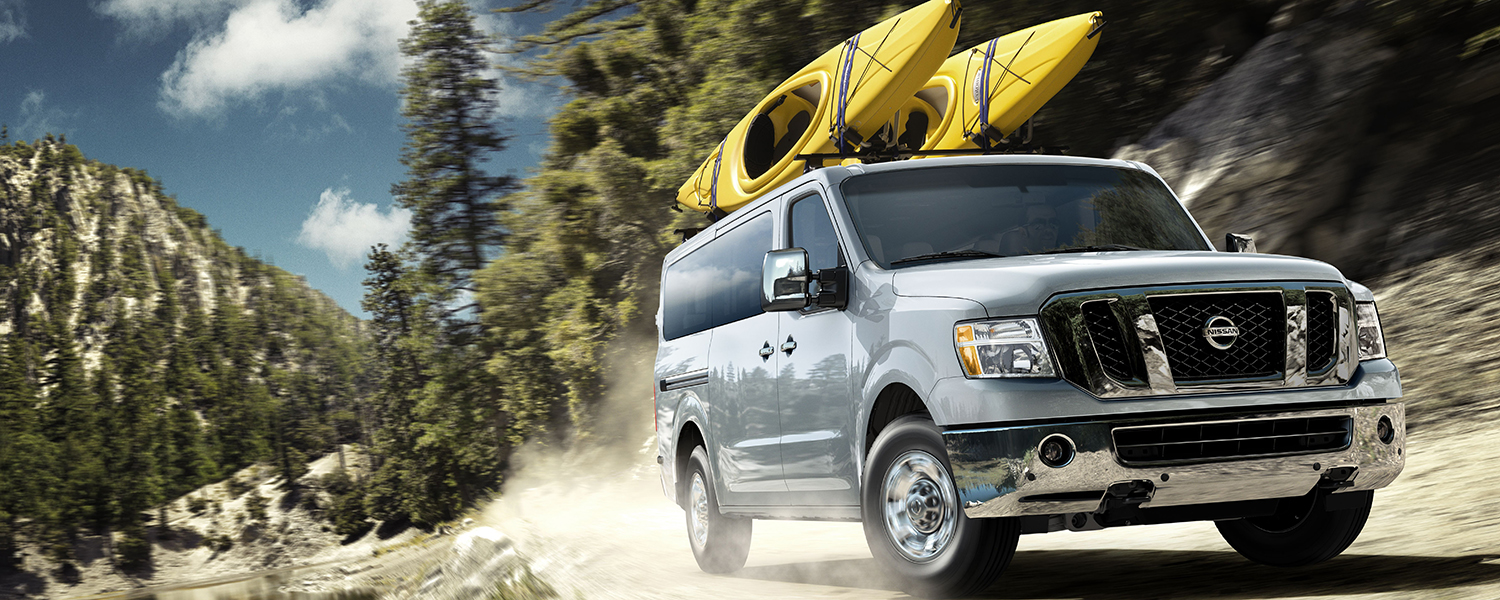 A silver NV Passenger van with kayaks loaded on the top, driving on a dirt road.