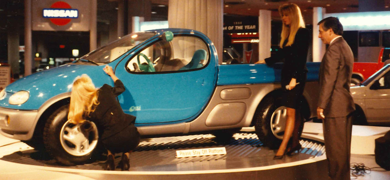 An old photo of 3 people looking at a light blue Nissan car.