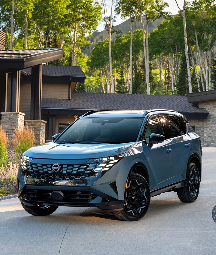 Front left 3/4 angle of the 2027 Nissan Rogue Hybrid Platinum parked in front of a house with trees behind.