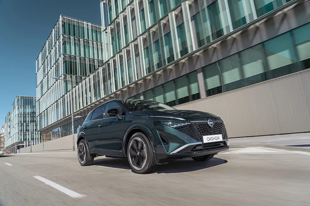 Green Nissan Qashqai driving on a road with a modern building in the background