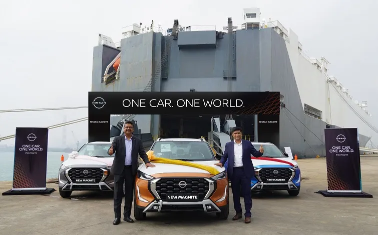 スーツを着た男性2人が新型日産マグナイトの前に立っています。背景にはインドから車を輸送する大型コンテナ船があり、他の車も2台見えます。