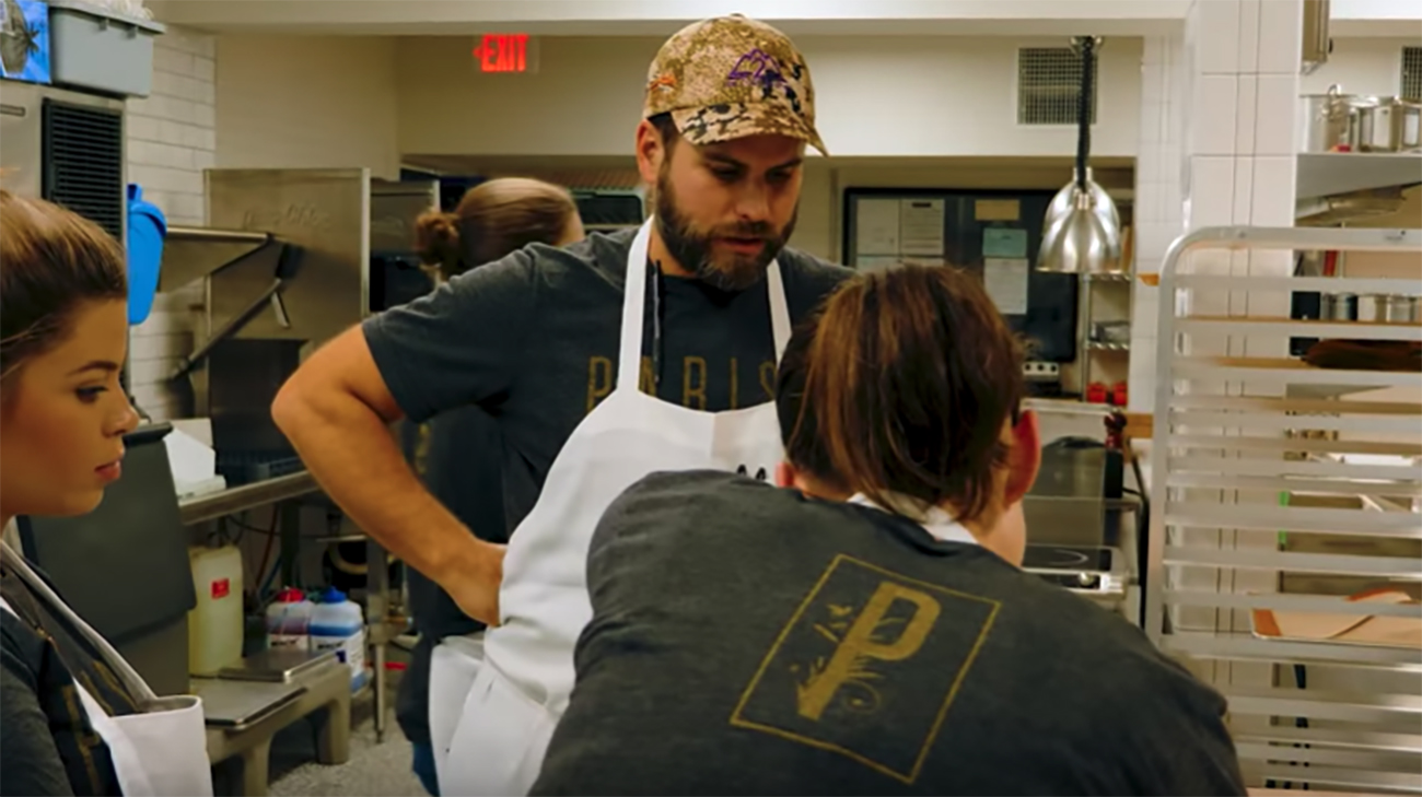 A man in white apron talking to his staff in a restaurant kitchen.