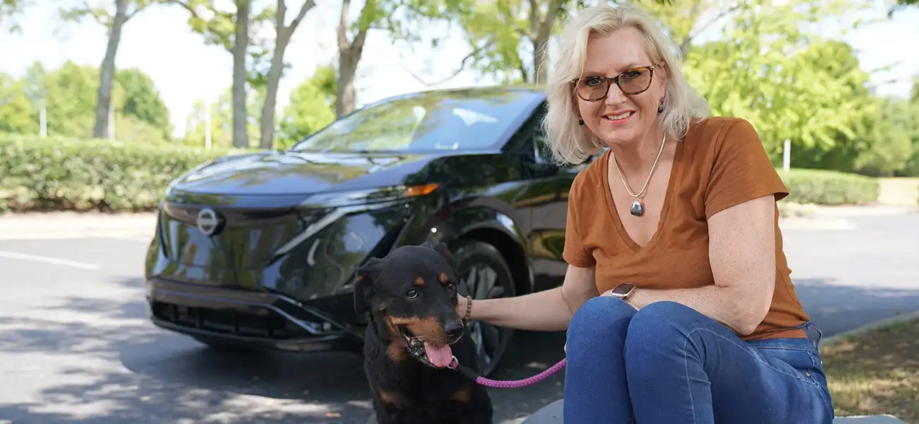 Nissan employee Henriëtte James sitting with her black and brown rescue dog, Tula, with a black Nissan Ariya and trees in the background