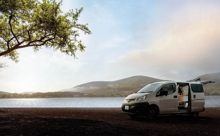 湖畔の木陰に停車している白いNV200バネット MYROOM。背景には湖と山並みが広がり、空は淡い雲に覆われている。車両はやや斜め前方を向いており、自然の中でアウトドア感を演出している。