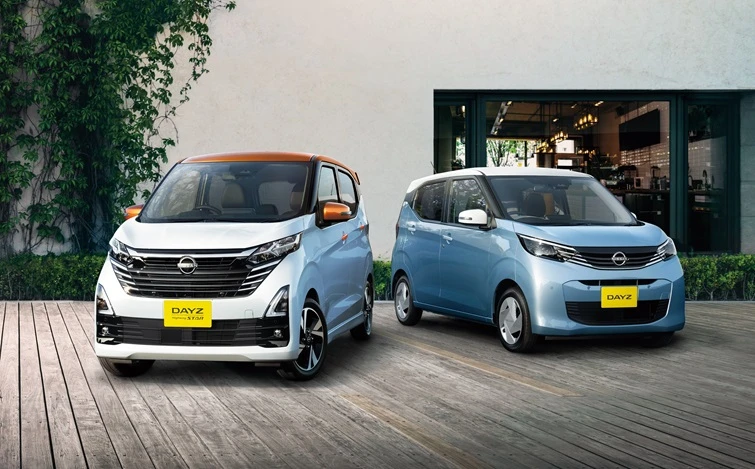 白いボディと水色のボディ、２台の軽自動車を正面から捉えたシーン。車両は建物のエントランス前に停車しており、周囲には緑豊かな樹木や壁面緑化が広がる。開放感のある屋外空間が描かれている。