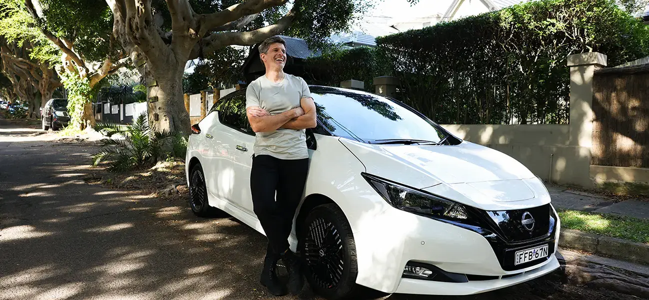 Nissan Australia ambassador Osher Gunsberg stands next to his white Nissan LEAF that is parked on a street