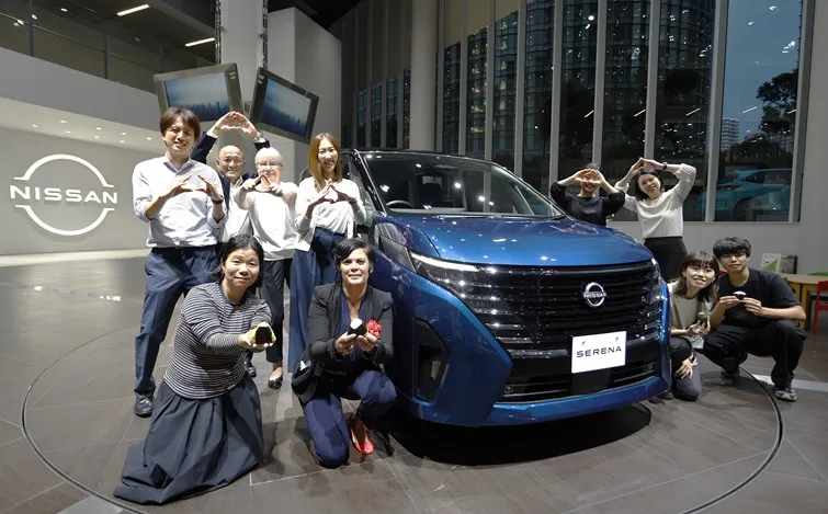 青い日産セレナがショールーム内に展示されている。車両は正面を向き、特徴的なフロントグリルとシャープなヘッドライトが際立っている。背景には大型ガラス窓越しに高層ビル群が見え、室内は明るい照明で近代的な雰囲気。車両の周囲には複数の人物が立っており、車の前でポーズを取っている様子がある。左側には「NISSAN」のロゴが壁面に表示されている。