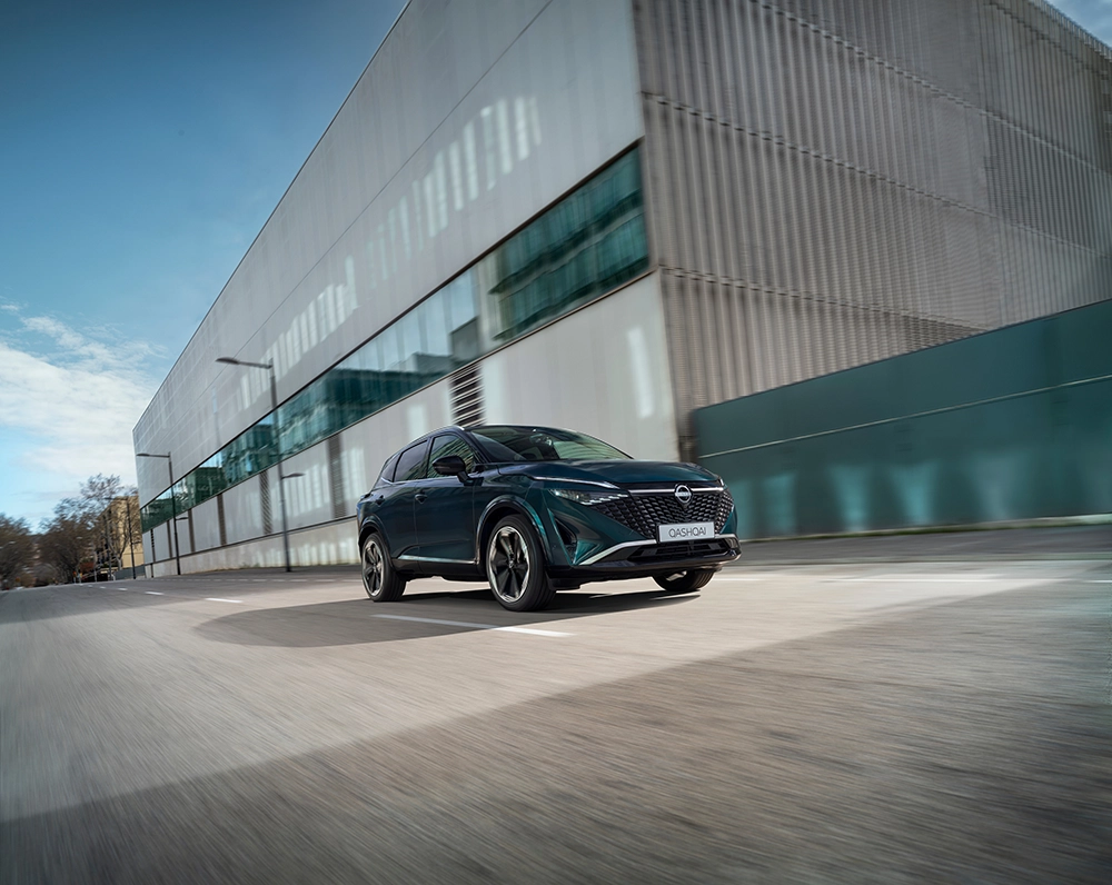 Green Nissan Qashqai driving on a road with a modern building in the background