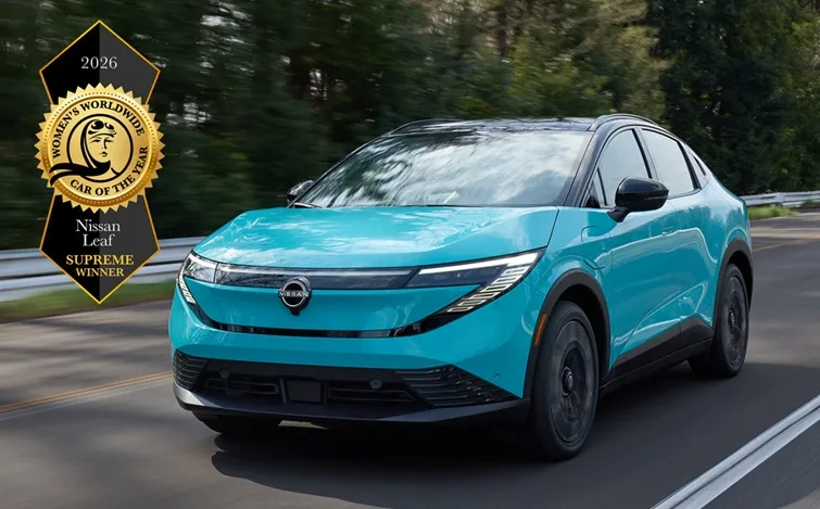 走行中の新型日産リーフが森林沿いの道路を前方へ進む様子を捉えた画像。左側に『2026 Women’s Worldwide Car of the Year Nissan Leaf Supreme Winner』の受賞ロゴが表示されている。