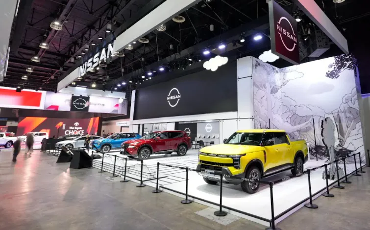 Wide view of a large Nissan booth at an indoor exhibition. Several vehicles are lined up facing forward on a white floor, with a yellow pickup truck positioned in the front right. Overhead lighting illuminates the space, and Nissan logos and display panels are visible in the background.