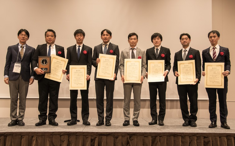 8人の人物が横一列に並び、賞状を持って立っている。背景は室内で、淡いベージュの壁と床が見える。人物は正面を向き、フォーマルな服装を着用。