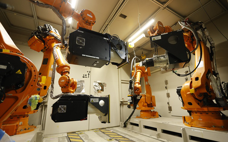 室内の作業エリアで、複数のオレンジ色の産業用ロボットアームが黒い装置を挟むように配置され、中央へ向けて稼働位置を取っている。背景は照明付きの天井と白い壁で、床には黄色と黒の注意ラインが引かれている。