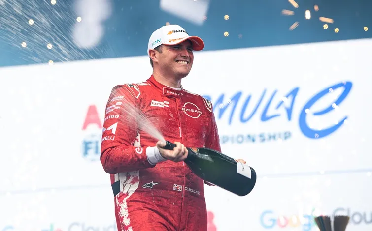サンパウロE-Prixで2位を獲得した日産フォーミュラEチームのオリバー ローランドが、赤いチームのレーススーツと白いキャップを着用し、表彰台でシャンパンを吹きかけている。
