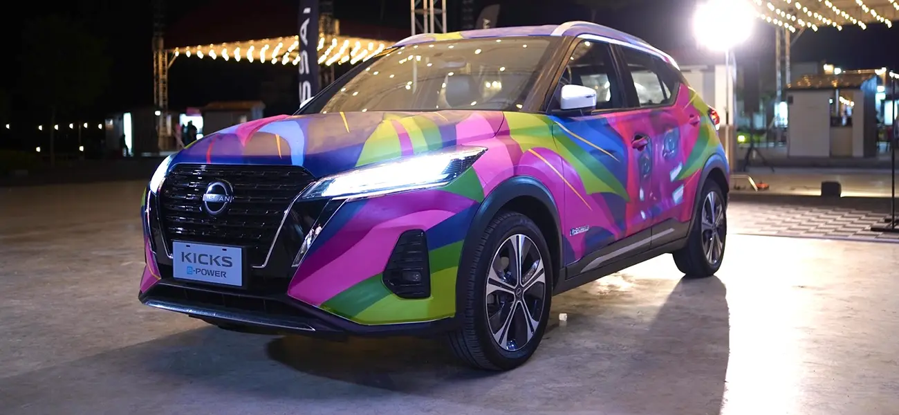 A Nissan Kicks e-POWER displayed outdoors at night, featuring a vibrant and colorful graffiti-style wrap with bright pink, green, yellow, and blue designs under event lighting.