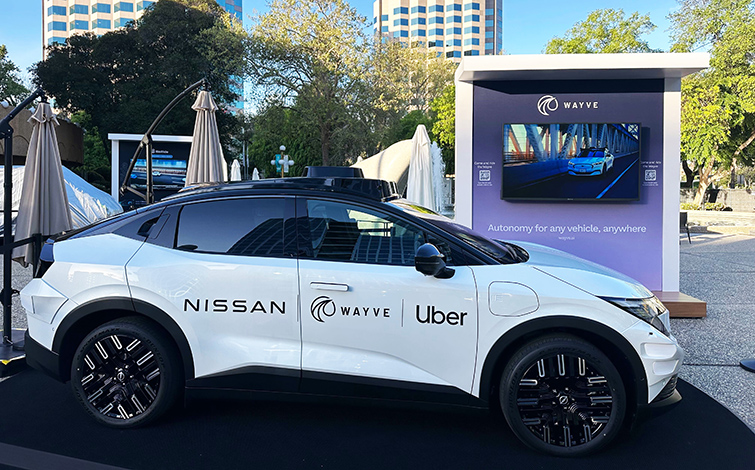 A white robotaxi prototype based on the new Nissan LEAF is shown from the side in an outdoor display area. Trees, tall buildings, and a booth with a large screen appear in the background.