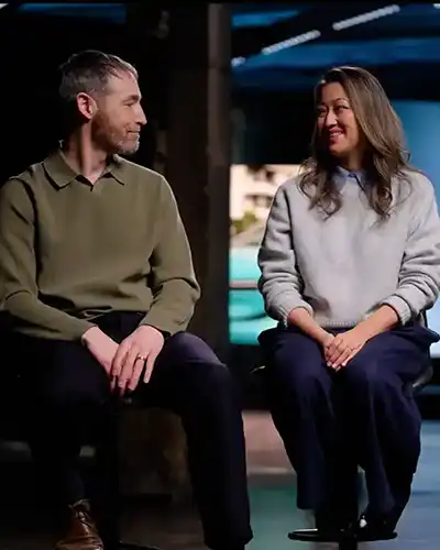 Chris und Julie Ramsey sitzen auf Hockern vor einem türkisfarbenen Auto und lächeln sich in einem dunklen Studio an.