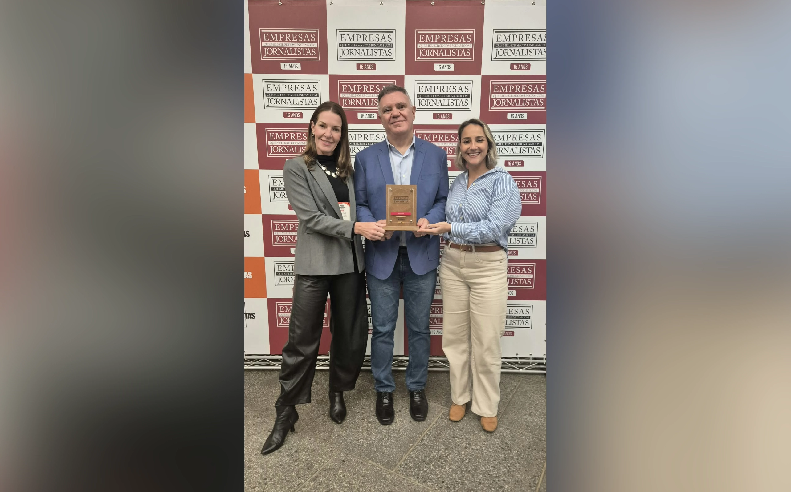 Três pessoas da equipe de Comunicação Corporativa da Nissan do Brasil seguram um troféu retangular à frente de um fundo quadriculado com a marca da Negócios da Comunicação