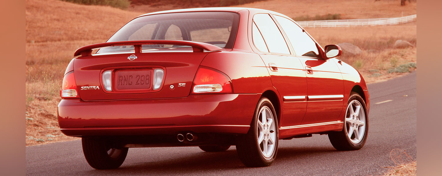 A red Nissan Sentra parked on a paved road with a white fence in the distance.