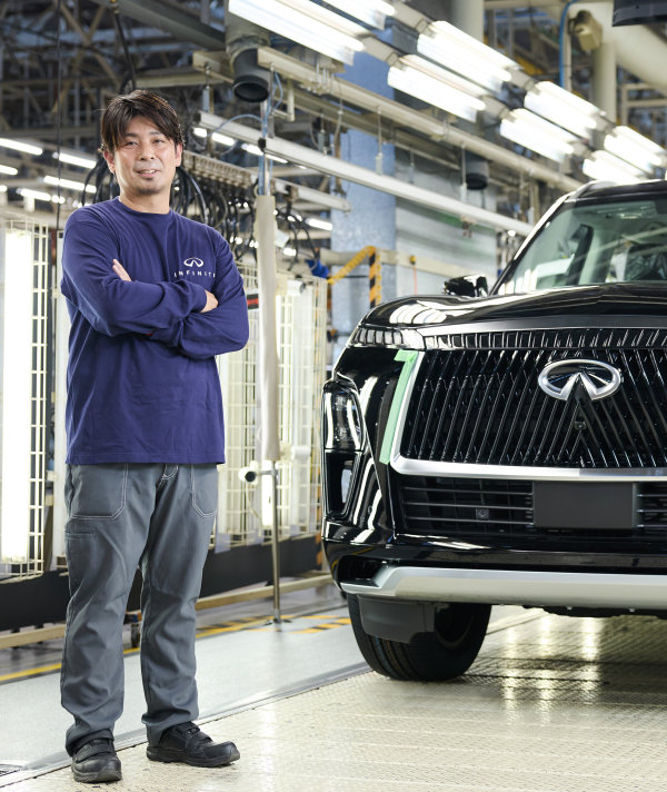 Akira Kiyota stands with his arms folded to the left of a black INFINITI QX80 inside the Nissan Shatai Kyushu plant.