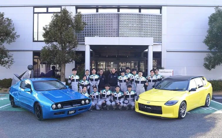 日産京都自動車大学校エントランスで左に青の「NEO SKYLINE」、右に黄色の「Z Lealia」を置いて集合写真に納まる学生と関係者たち
