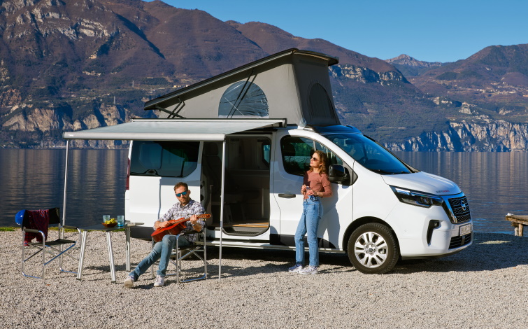 Seitenansicht des Primastar Campers mit aufgestelltem Dachzelt, Vorzelt und zwei Personen an einem See