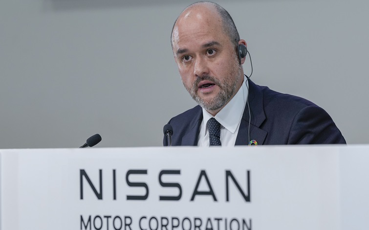 日産自動車 2025年度第1四半期決算発表の会場にて、スーツ姿の日産自動車CEO イヴァン エスピノーサが「NISSAN MOTOR CORPORATION」と記された演壇に着席している。背景はグレー。