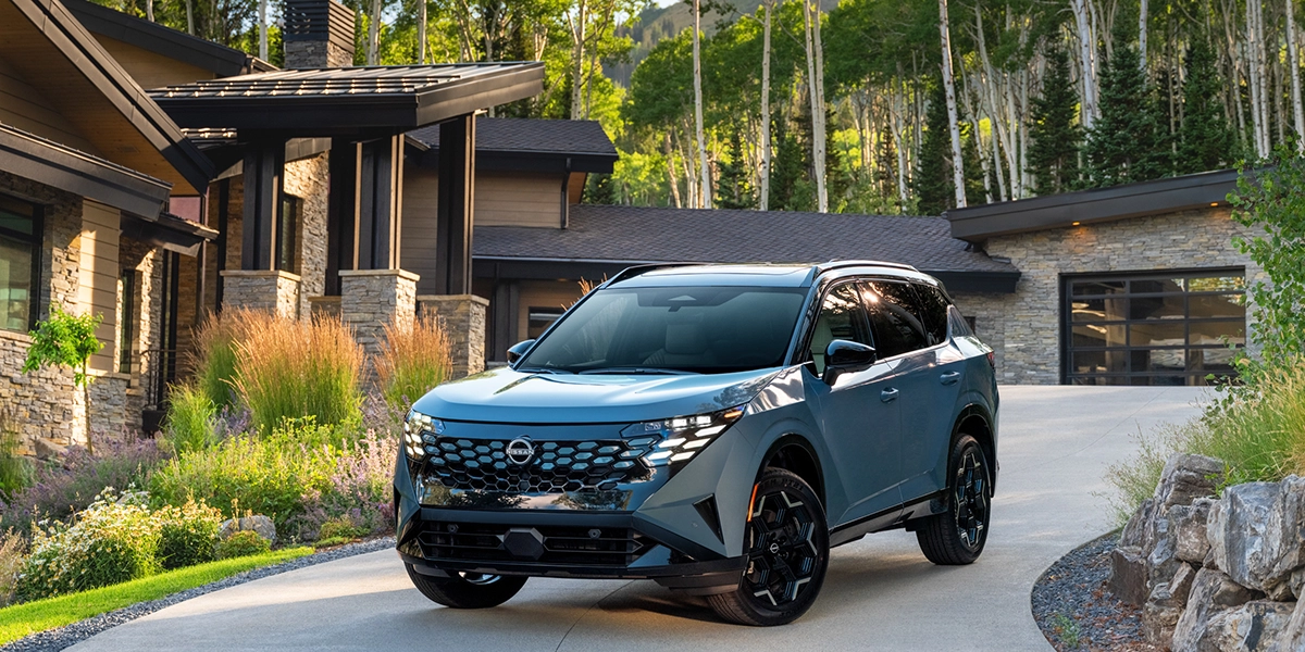 Front left 3/4 angle of the 2027 Nissan Rogue Hybrid Platinum parked in front of a house with trees behind.