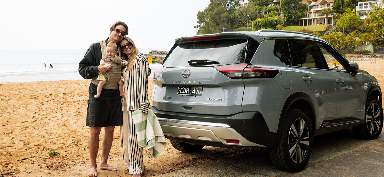 A man and a woman holding a baby, standing on a beach in front of their parked Nissan X-Trail