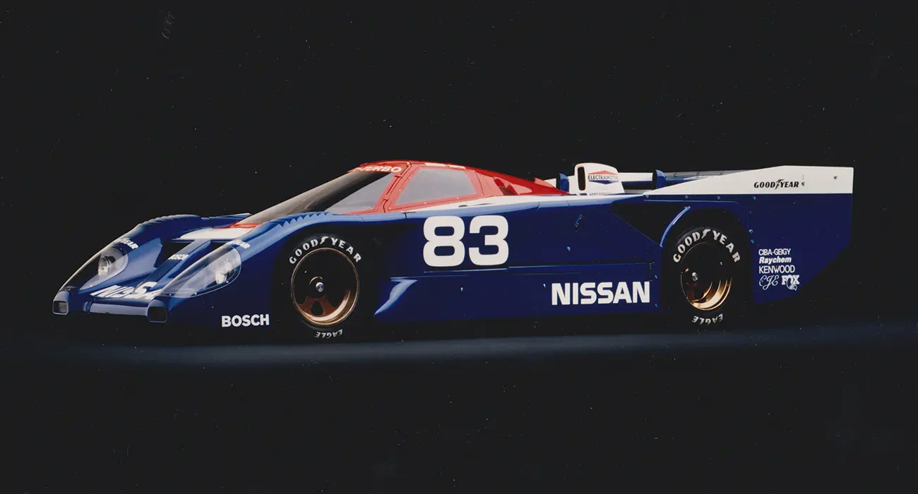 Front left 3/4 angle of a blue, white, and red Nissan racing car against a black bag round with shadowing.