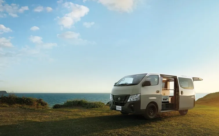 海沿いの草地に停車したキャニオンベージュのキャラバンMYROOM。右側面とフロントが見える角度で、リアゲートを開けている。背景には青空と海の水平線が広がり、柔らかな日差しが車体に当たっている。