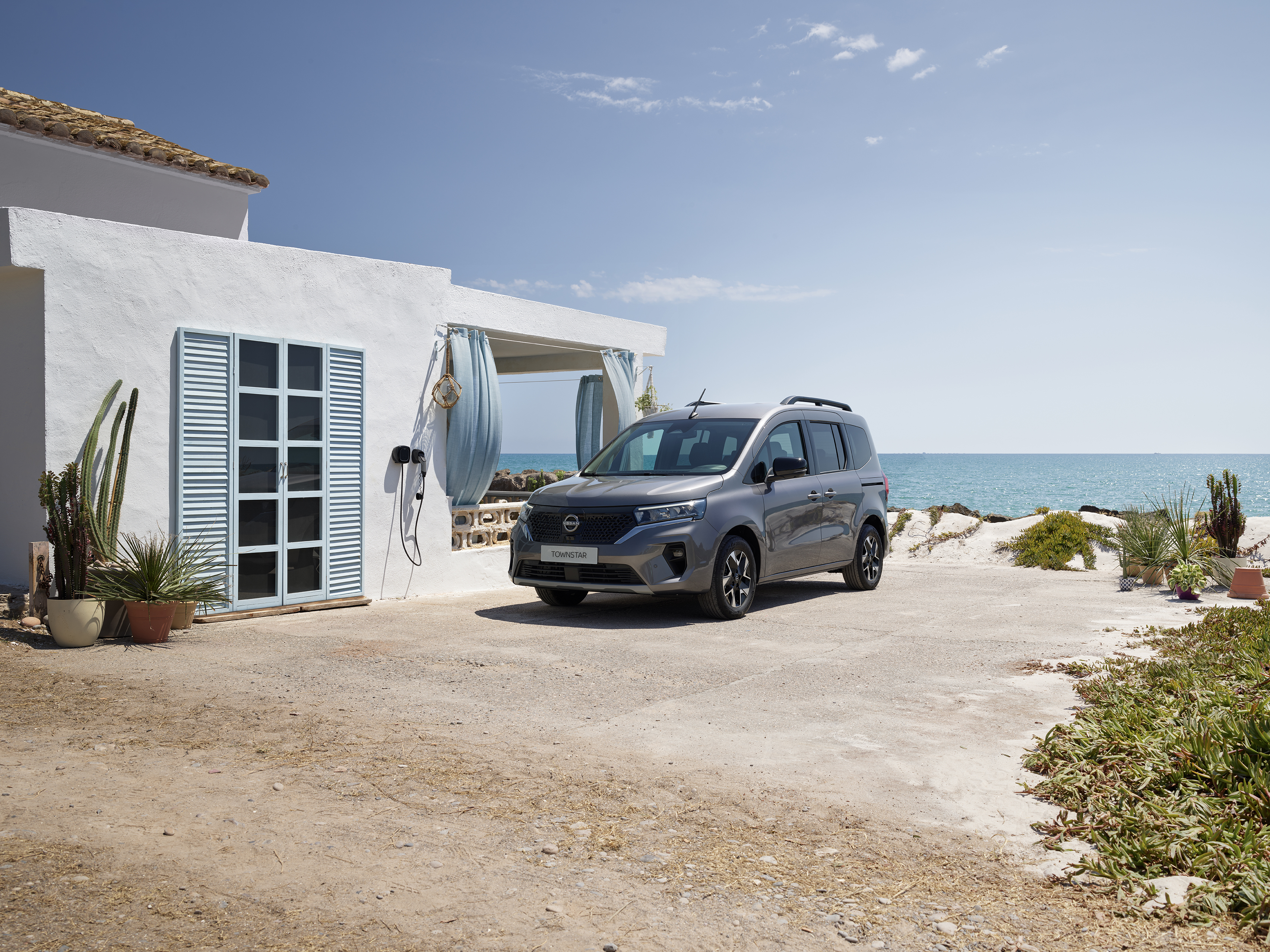3/4 Frontansicht eines grauen Nissan Townstar EV Kombi, der an einem hellen Haus steht, im Hintergrund das Meer