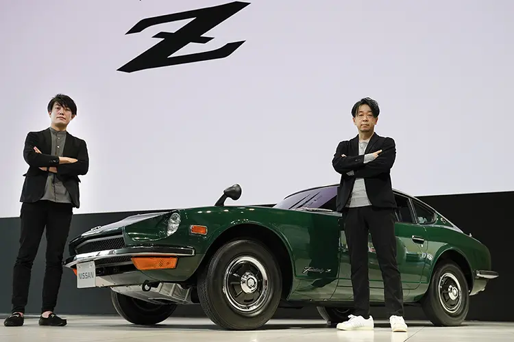 Two people standing on stage facing forward with arms crossed in front of green car