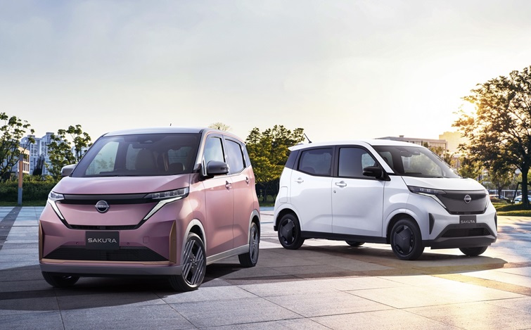 青空の広がる屋外の舗装された広場で、ピンク色の「日産サクラ」が手前左から斜め前向きに走行しており、背景奥には白い日産サクラが同じ方向に並んで走っている様子を捉えた画像。周囲には街路樹が見え、開放的な都市空間が広がっている。