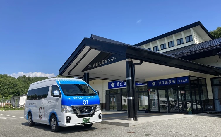 Under a clear blue sky, the Nissan Caravan for No.1 car of "Namie Smart Mobility" is parked facing left-front in front of the Namie Town Office Tsushima Branch. The white and blue vehicle stands in the foreground, with the building, paved area, and green trees visible in the background.