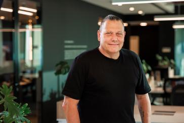 Guy Rodríguez, homem branco e cabelos castanhos e presos, de camiseta cinza olhando para a câmera apoiado em uma mesa.