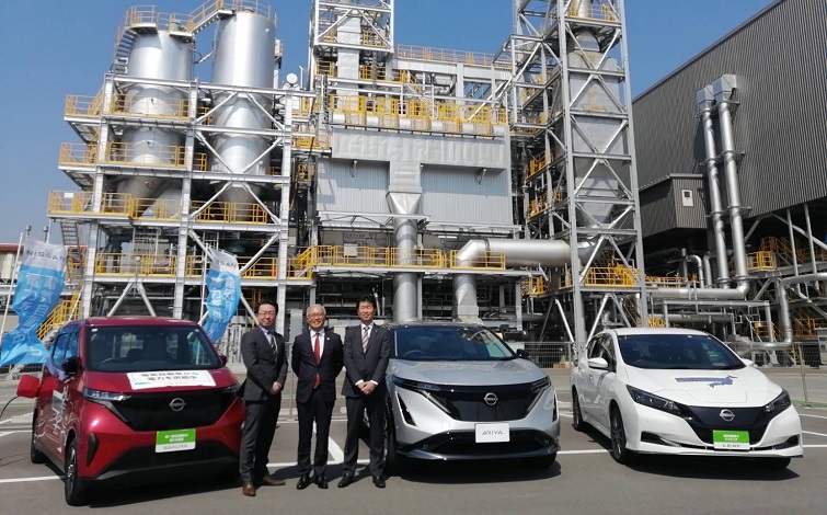 電気自動車（サクラ、アリア、リーフ）の前で記念撮影する各代表者（スーツ姿の男性３名。背景はプラント）