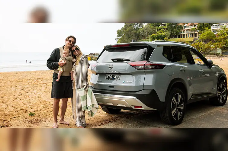 A man and a woman holding a baby, standing on a beach in front of their parked Nissan X-Trail